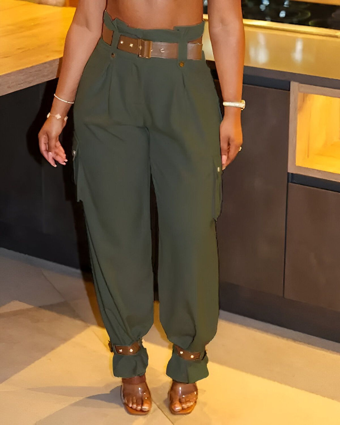 Une femme porte un pantalon cargo vert olive taille haute avec une ceinture marron. Elle porte également des sandales à talons transparentes et un haut court noir.
