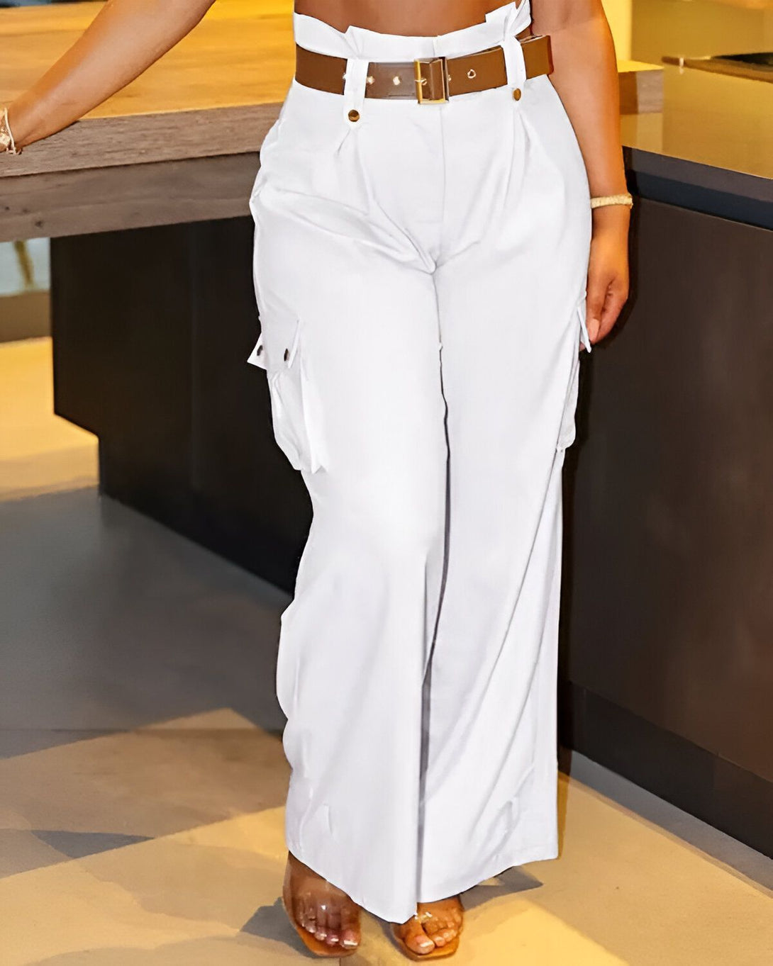 Une femme porte un pantalon cargo blanc taille haute avec une ceinture marron. Le pantalon est ample et a des poches cargo sur les côtés. La femme est debout dans une cuisine et est vue de face.