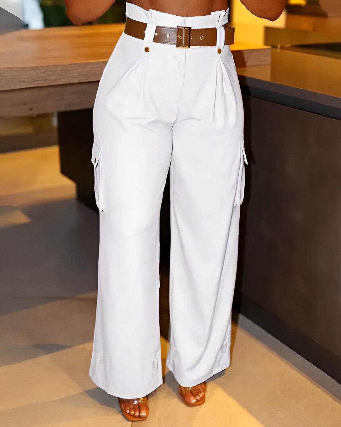 Une femme porte un pantalon cargo blanc taille haute avec une ceinture marron. Elle est debout dans une cuisine et porte des sandales marron clair.