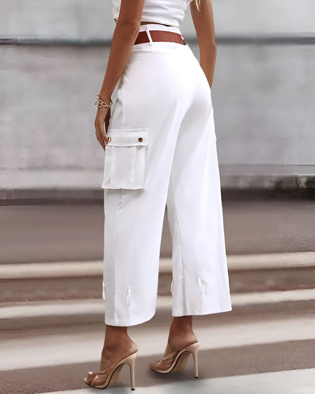 Une femme porte un pantalon cargo blanc taille haute avec des poches sur les côtés. Le pantalon est ample et évasé. Elle porte également des talons hauts transparents.