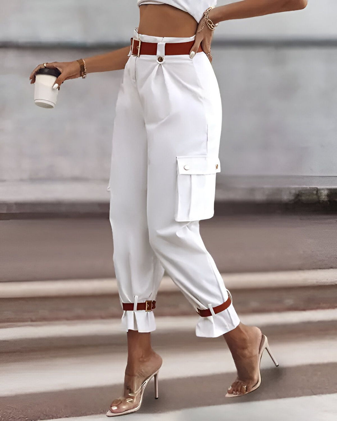 Une femme porte un pantalon cargo blanc avec une ceinture marron et des talons hauts transparents. Elle tient une tasse de café à la main.
