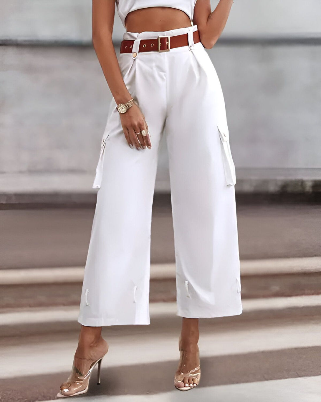 Une femme porte un pantalon cargo blanc taille haute avec une ceinture marron. Elle porte également des talons transparents et un haut court blanc.
