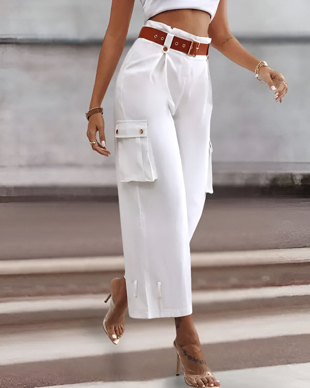 Une femme porte un pantalon cargo blanc taille haute avec une ceinture marron. Elle porte également un crop top blanc et des sandales à talons transparentes.