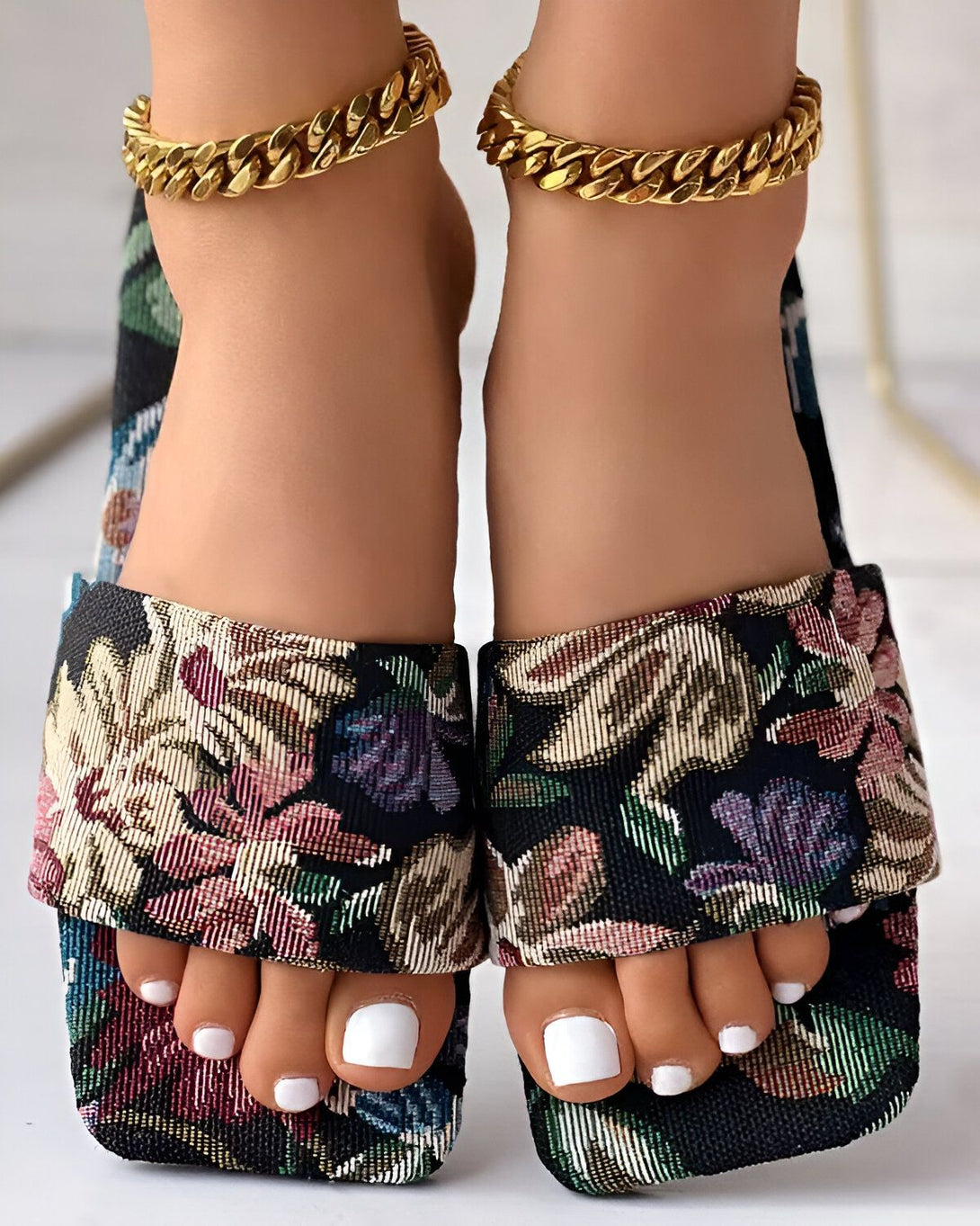 Une femme porte des sandales à talons hauts à bout carré avec un motif floral coloré. Les sandales sont dotées d'une bride de cheville dorée.