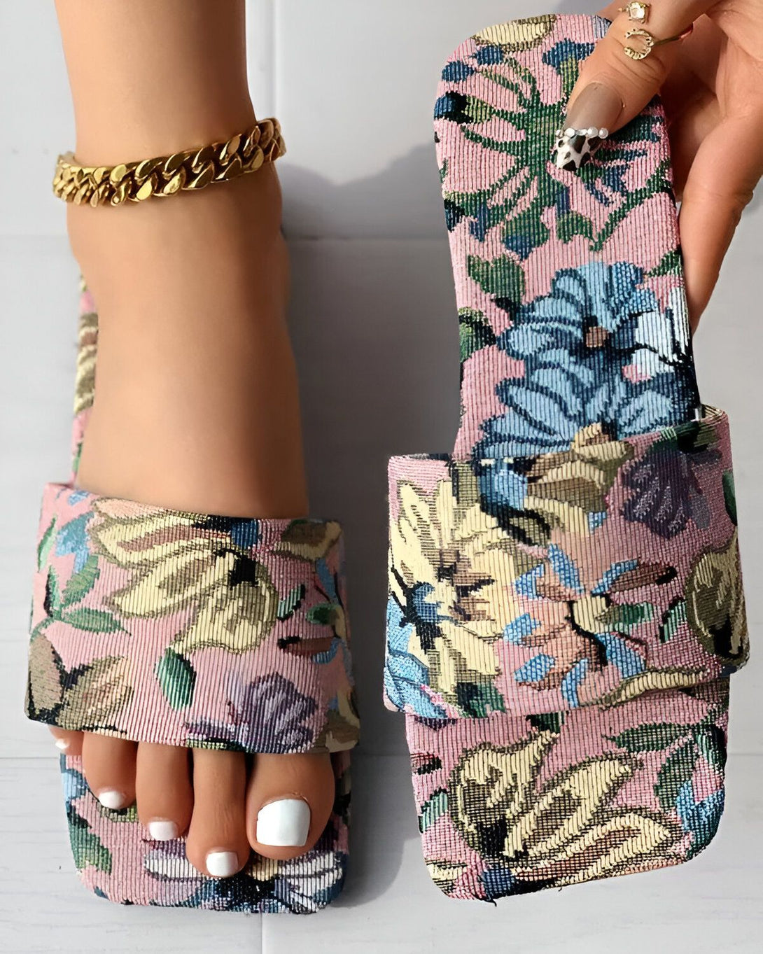 Une femme porte des sandales plates à bout carré roses à motif floral. Les sandales sont en tissu et ont une semelle intérieure légèrement rembourrée. La femme a les ongles des pieds peints en blanc et porte un bracelet de cheville en or.
