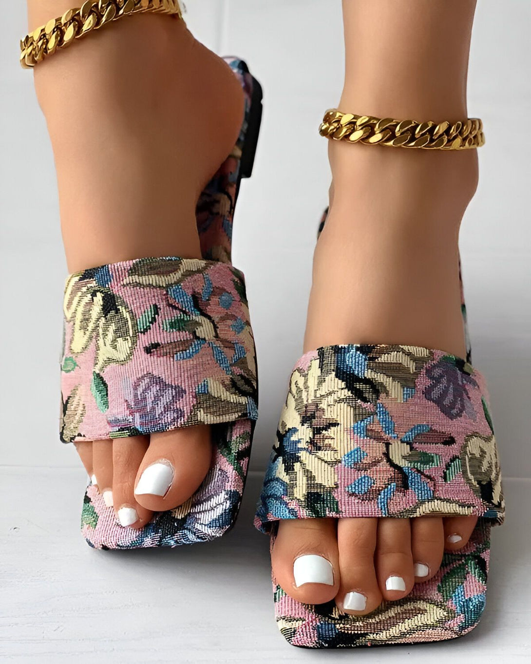 Une femme porte des sandales à talons hauts à bout carré. Les sandales sont roses et comportent un motif floral. La femme a les ongles des pieds peints en blanc et porte un bracelet de cheville en or.