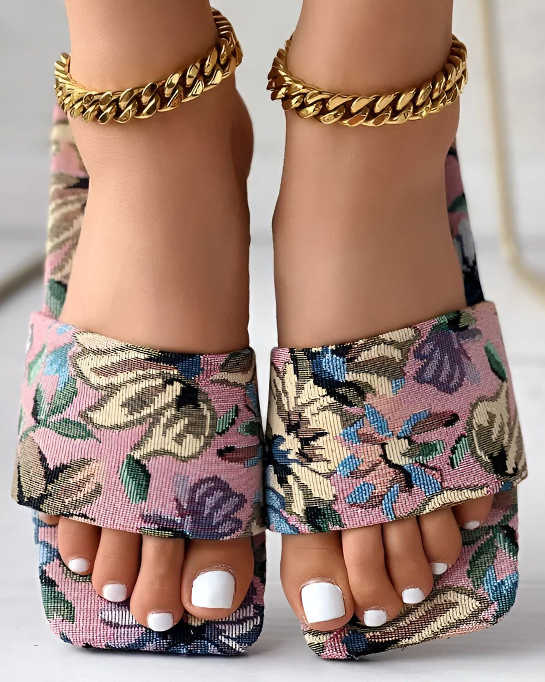 Une femme porte des mules à talons hauts à bout carré. Les mules sont en tissu rose à motifs floraux et ont une semelle intérieure blanche. La femme a les ongles des pieds peints en blanc et porte des bracelets de cheville en or.