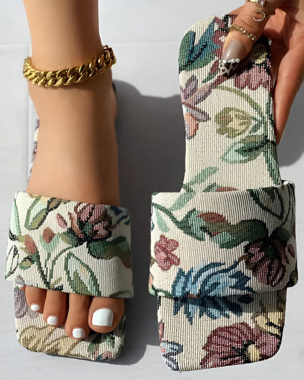 Une femme porte des sandales plates à enfiler. Les sandales sont blanches avec un motif floral coloré. La femme a les ongles des pieds peints en blanc et porte un bracelet de cheville en or.