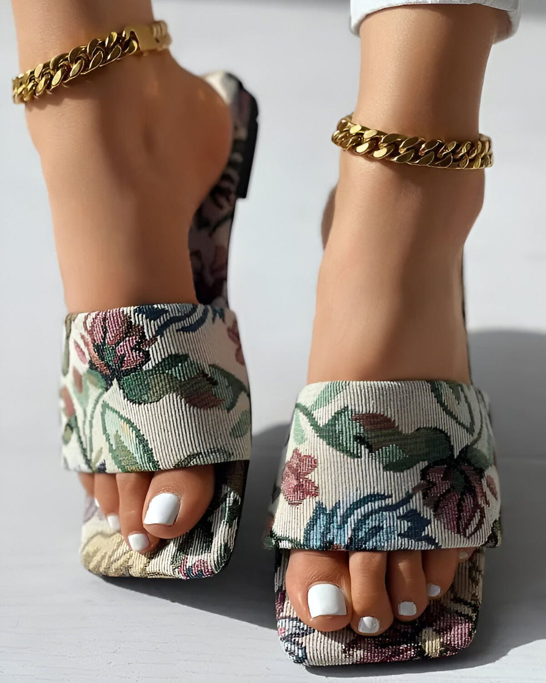 Une femme porte des sandales à talons hauts à bout carré avec un motif floral et des brides dorées. Elle porte également un bracelet de cheville en or.