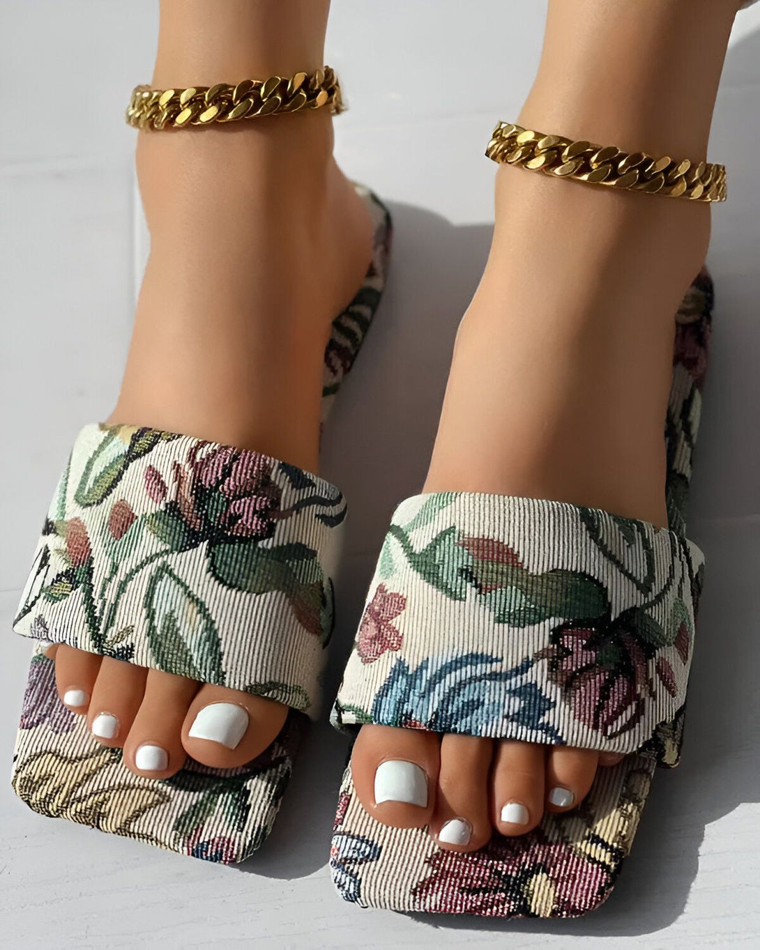 Une femme porte des sandales plates à enfiler avec un motif floral coloré et des brides dorées.