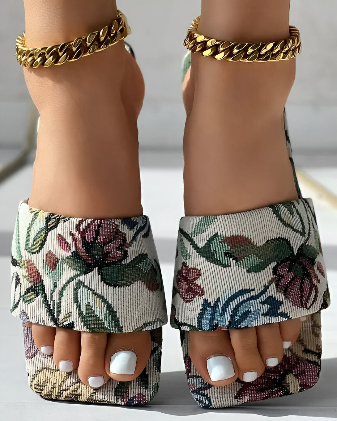 Une femme porte des sandales à talons hauts à bout carré avec un motif floral coloré. Les sandales sont dotées d'une bride de cheville dorée.