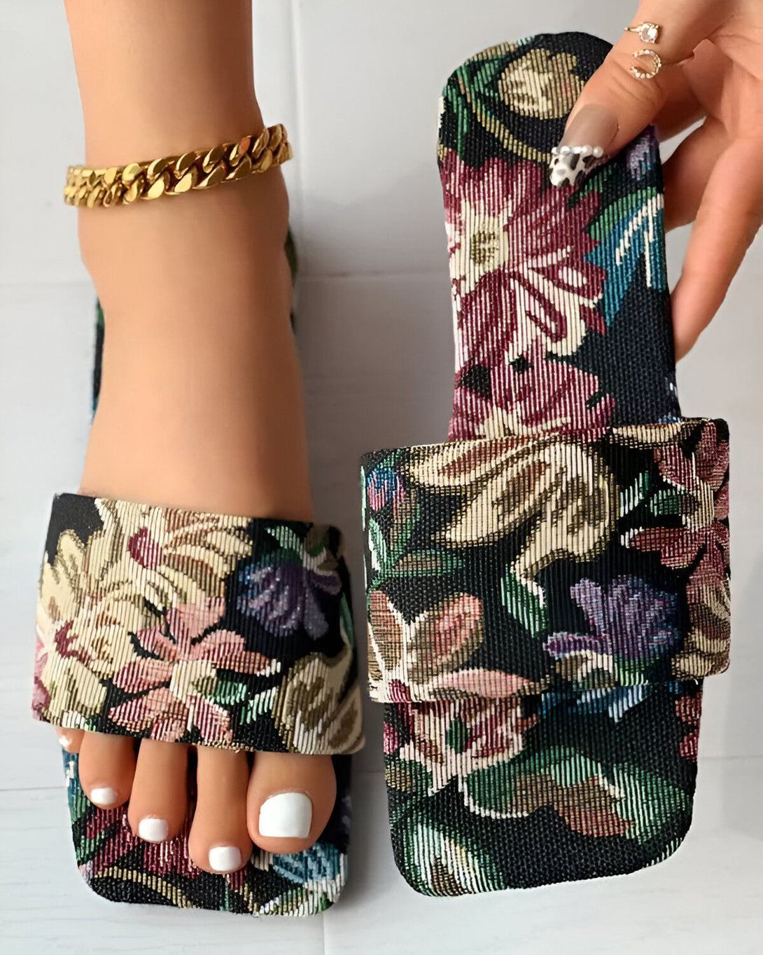 Une femme porte des sandales plates à bout carré avec un motif floral coloré. Les sandales sont en tissu et ont une semelle intérieure rembourrée. La femme a les ongles des pieds peints en blanc et porte un bracelet de cheville en or.