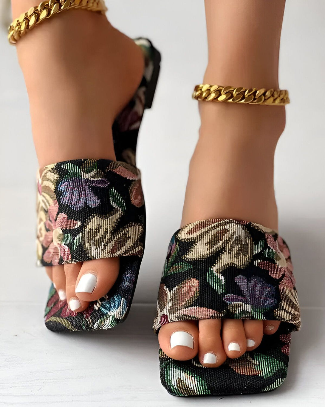 Une femme porte des sandales à talons hauts à bout carré. Les sandales sont noires avec un motif floral coloré. La femme a les ongles des pieds peints en blanc. Elle porte également un bracelet de cheville en or.