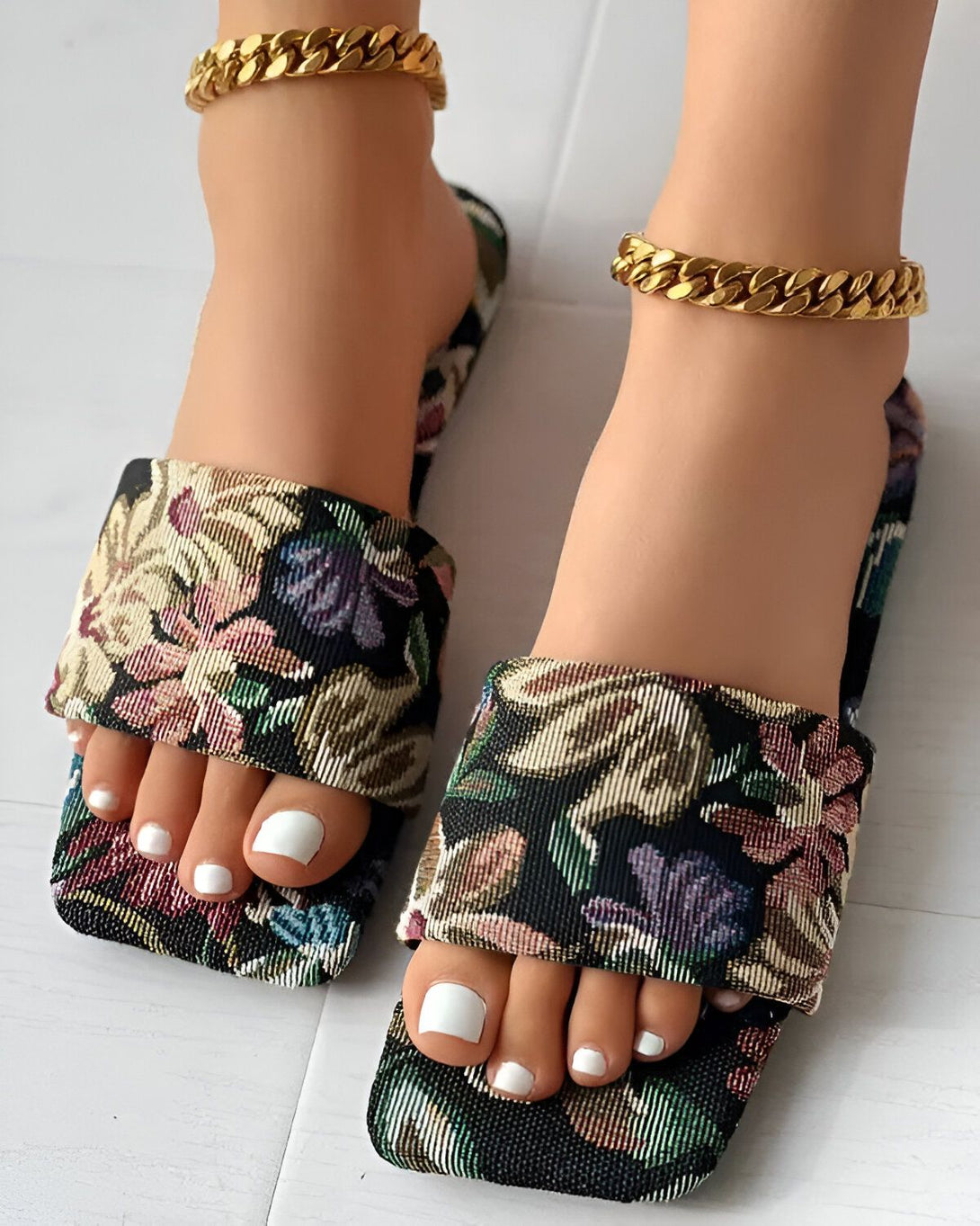 Une paire de sandales plates à enfiler avec un motif floral coloré et une semelle intérieure rembourrée pour plus de confort. Les sandales sont portées par une femme avec un pantalon blanc et un haut noir.