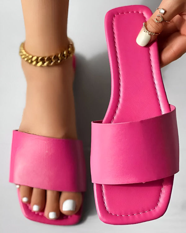 Une femme porte des sandales roses. Les sandales sont en cuir et ont une semelle plate. La femme a les ongles des pieds peints en blanc et porte un bracelet de cheville en or.