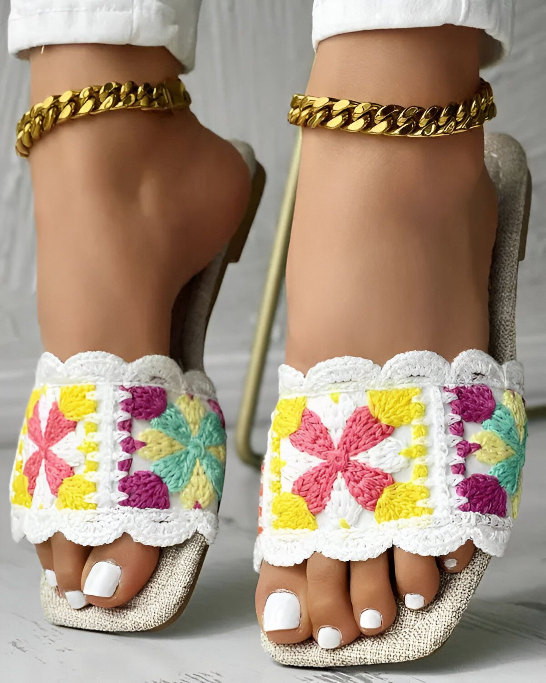 Une femme porte des sandales plates blanches avec des fleurs colorées brodées dessus. Elle porte également une chaîne en or autour de la cheville.