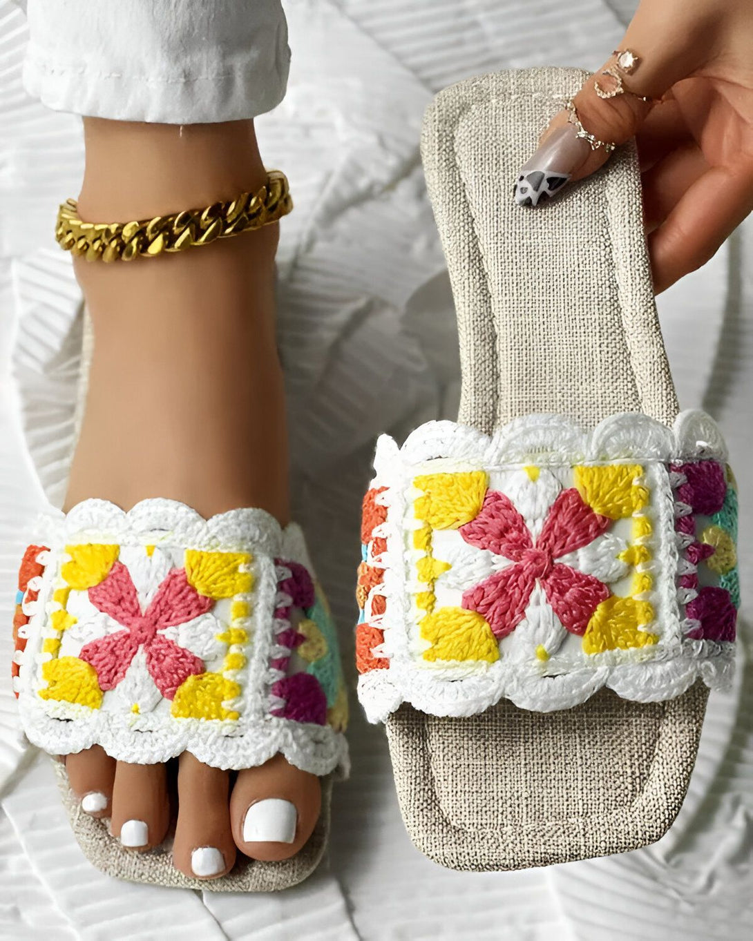 Une femme porte des sandales plates blanches avec des fleurs colorées brodées sur le dessus. Les sandales sont en crochet et ont une semelle en jute. La femme porte également un pantalon blanc et un haut blanc. Elle est assise sur une chaise et tient les sandales dans sa main.
