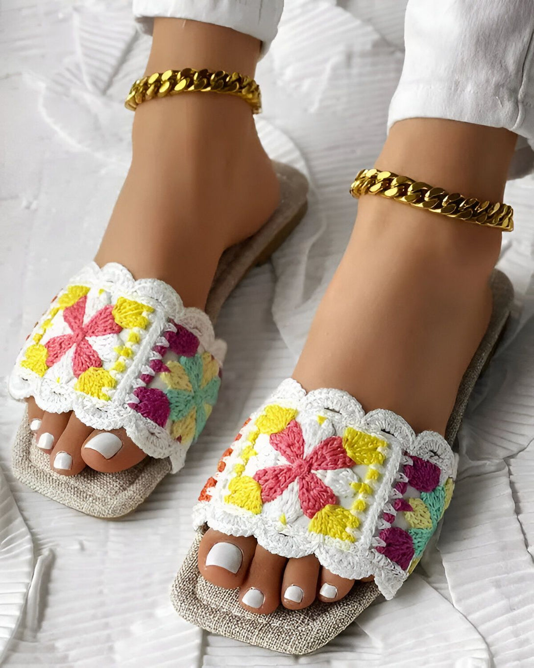 Une paire de sandales plates blanches avec des fleurs colorées brodées dessus. Les sandales sont portées par une femme qui a les ongles des pieds peints en blanc. La femme porte également un pantalon blanc et un bracelet de cheville doré.