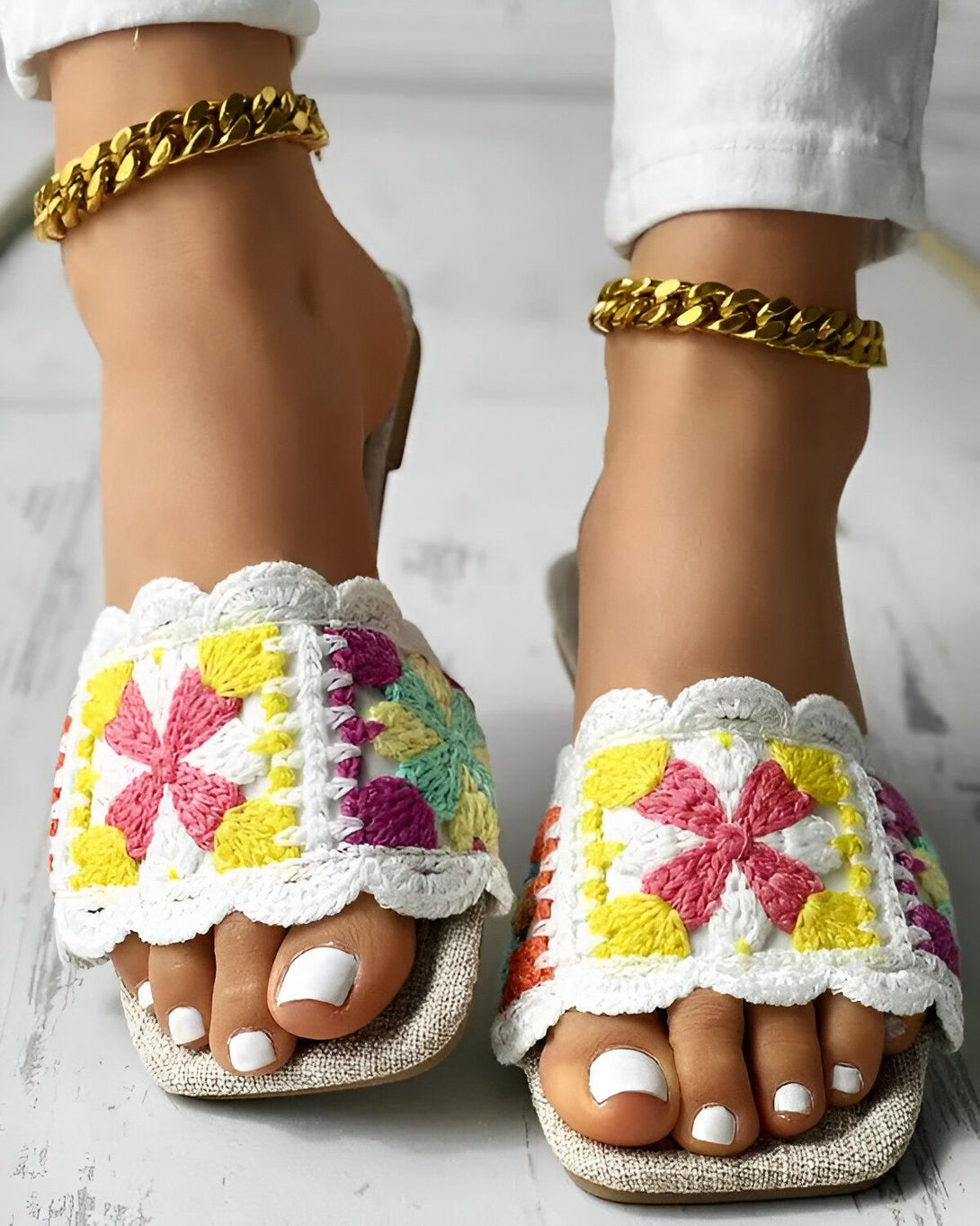 Une femme porte des sandales plates blanches avec des fleurs colorées brodées dessus. Elle porte également un pantalon blanc et un bracelet de cheville doré.