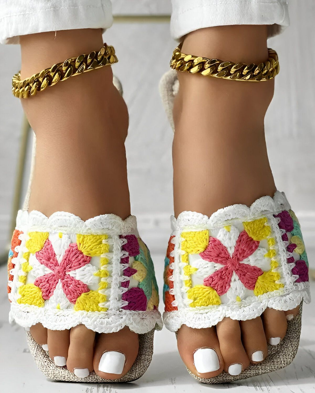Une femme porte des sandales colorées avec des motifs floraux. Les sandales sont faites de fil et ont une semelle en corde. La femme a les ongles des pieds peints en blanc et porte des bracelets de cheville en or.
