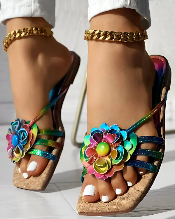 Une femme porte des sandales plates à fleurs multicolores. Les sandales ont des brides fines et une semelle compensée en liège. La femme a les ongles des pieds peints en blanc et porte un bracelet de cheville doré.