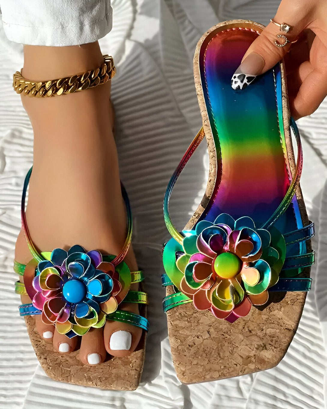 Une femme porte des sandales à talons compensés arc-en-ciel avec des fleurs en métal colorées. Les sandales sont en liège et ont une semelle antidérapante. La femme a les ongles des pieds peints en blanc et porte un bracelet de cheville en or.