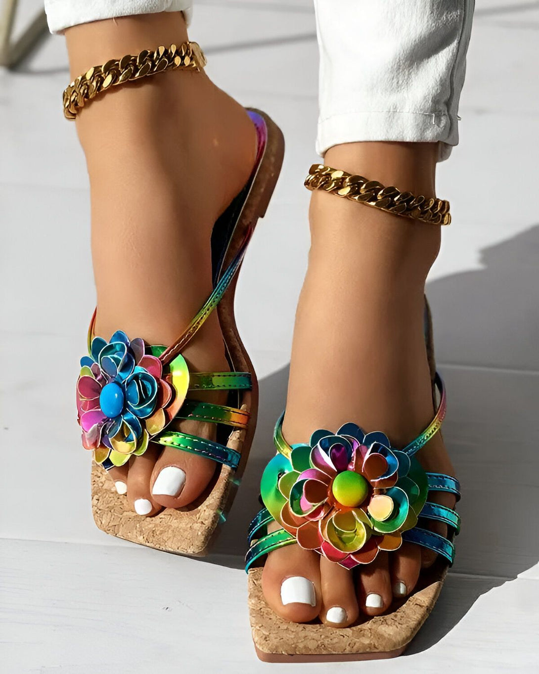 Une femme porte des sandales plates à fleurs multicolores. Les sandales ont des brides fines et une semelle en liège. La femme a les ongles des pieds peints en blanc et porte un pantalon blanc et une cheville dorée.