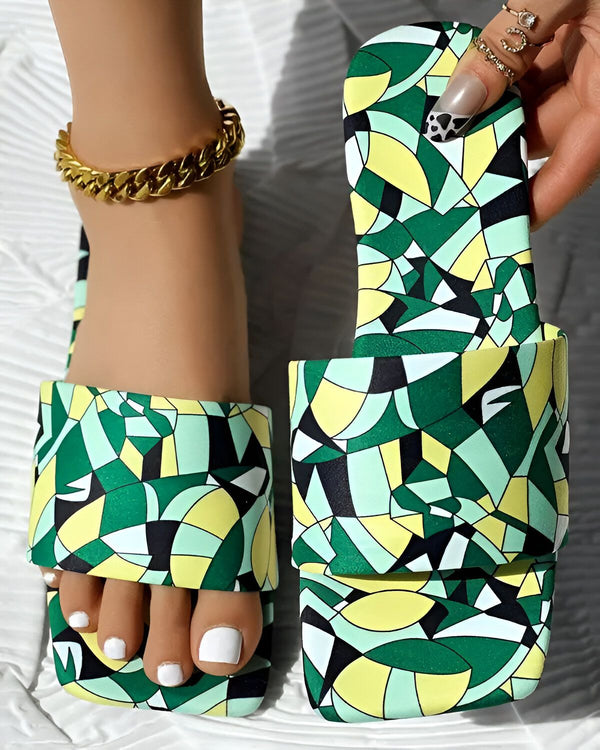Une femme porte des sandales colorées avec un motif géométrique. Les sandales sont vertes, jaunes et noires. La femme a les ongles des pieds peints en blanc et porte un bracelet de cheville doré.