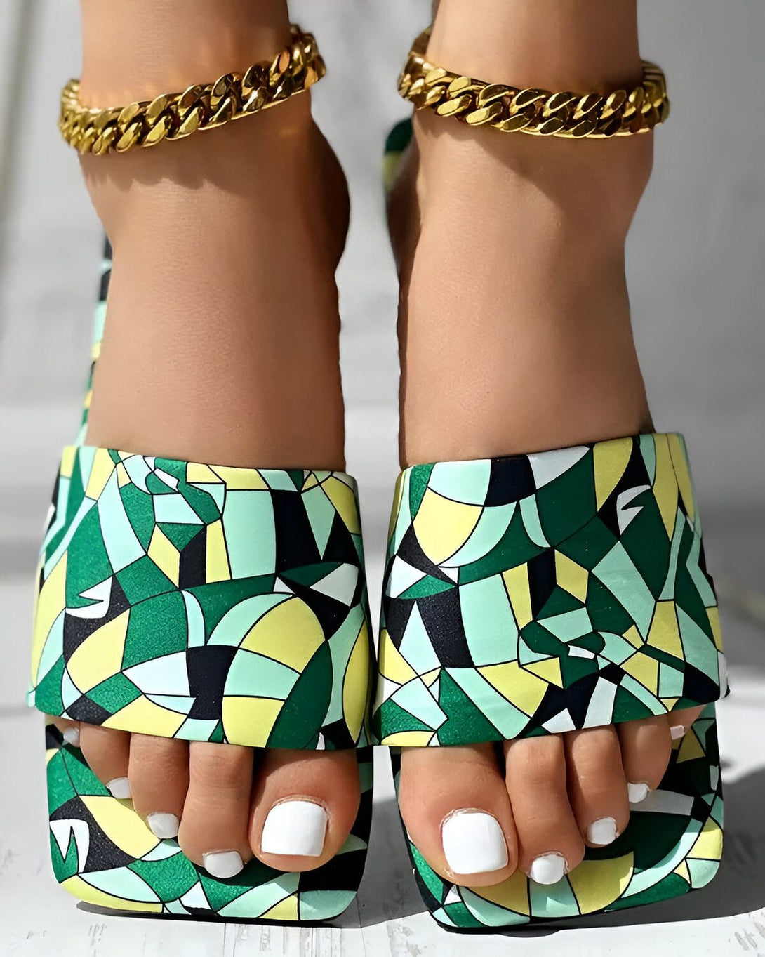 Une femme porte des sandales à talons hauts multicolores. Les sandales sont à bout ouvert et ont une bride qui s'enroule autour de la cheville. La femme a les ongles des pieds peints en blanc et porte des bracelets de cheville en or.
