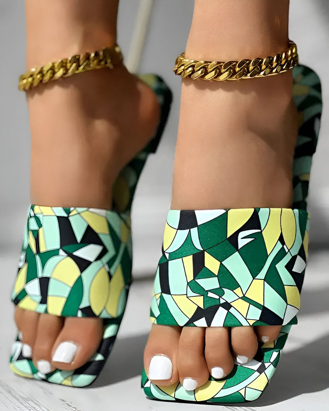 Une femme porte des sandales colorées à talons hauts avec un motif géométrique vert, jaune et noir. Elle porte également une chaîne de cheville en or.