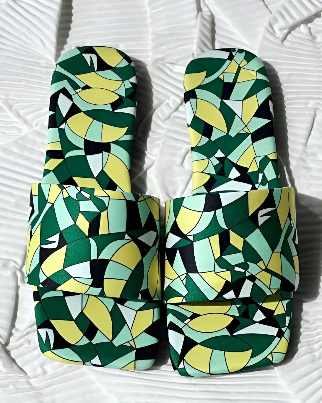 Une paire de sandales plates à enfiler, avec un motif géométrique coloré. Les sandales sont jaune, vert et noir.