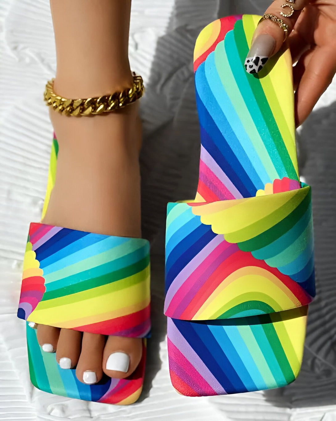 Une femme porte des sandales plates à enfiler. Les sandales sont colorées et ont un motif arc-en-ciel. La femme a les ongles peints en noir et porte un bracelet de cheville doré.