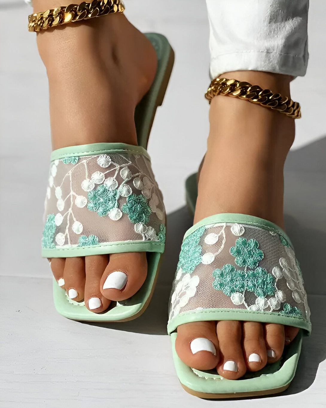 Une femme porte des sandales plates vertes avec des fleurs brodées. Les sandales sont ouvertes au niveau du talon et ont une bride qui passe entre les orteils. La femme a les ongles des pieds peints en blanc et porte des bijoux de cheville en or.