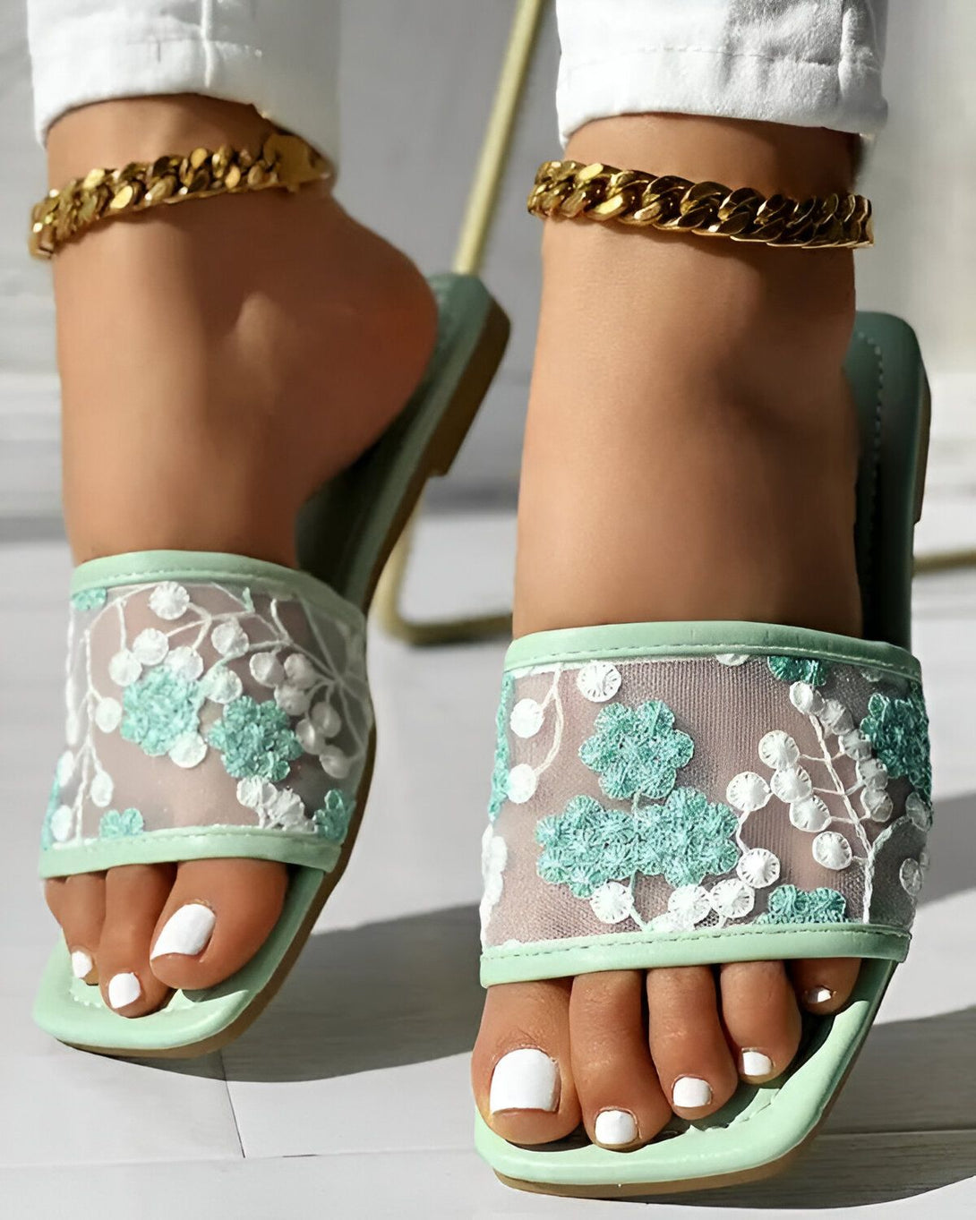 Une femme porte des sandales plates vertes à bout carré avec un motif floral brodé sur le dessus. Les sandales sont en mesh et ont une semelle intérieure légèrement rembourrée. La femme a les ongles des pieds peints en blanc et porte un bracelet de cheville en or.