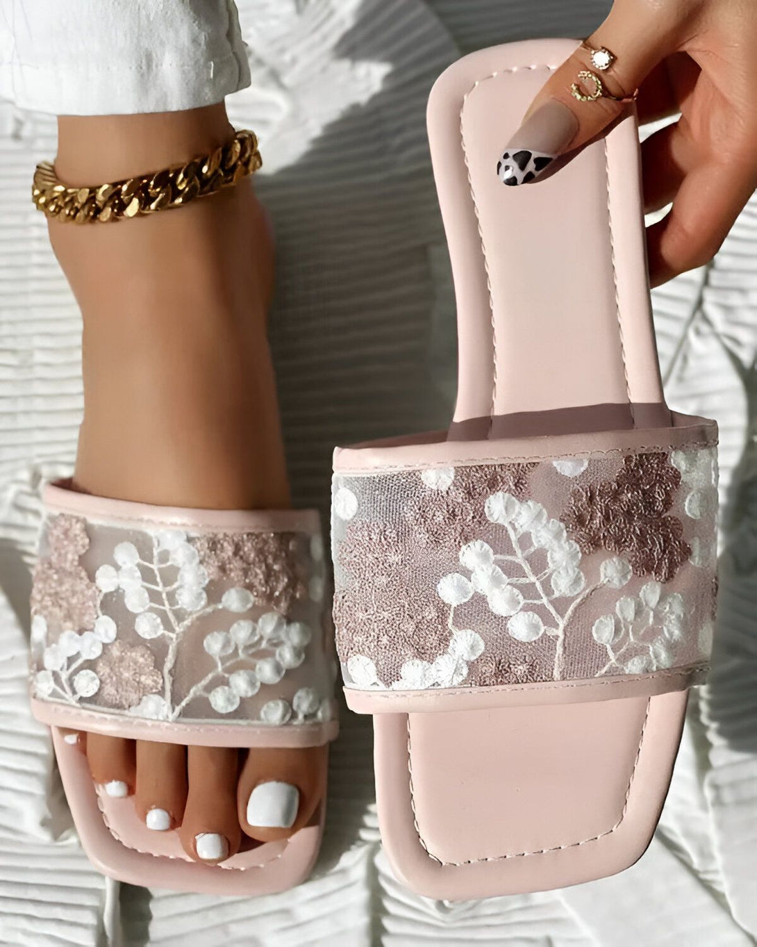 Une femme porte des sandales roses à enfiler. Les sandales sont en dentelle et ont un bout carré. La femme a les ongles des pieds peints en blanc et porte un bracelet de cheville en or.