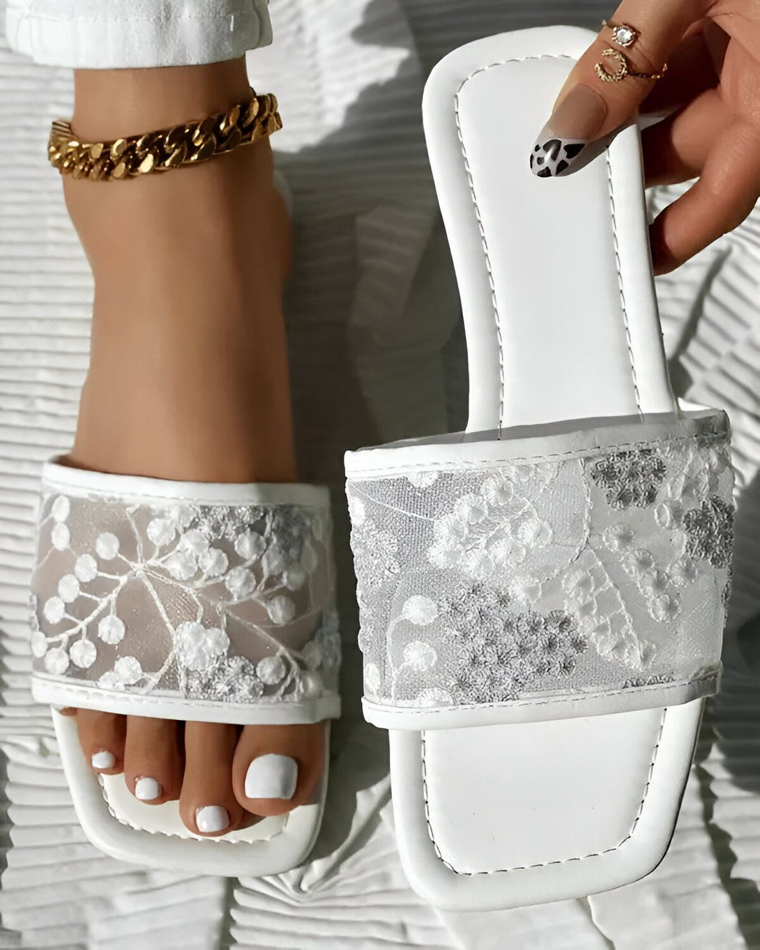 Une femme porte des sandales blanches à talons hauts. Les sandales sont en dentelle et ont un bout ouvert. La femme a les ongles des pieds peints en blanc. Elle porte également un bracelet de cheville en or.