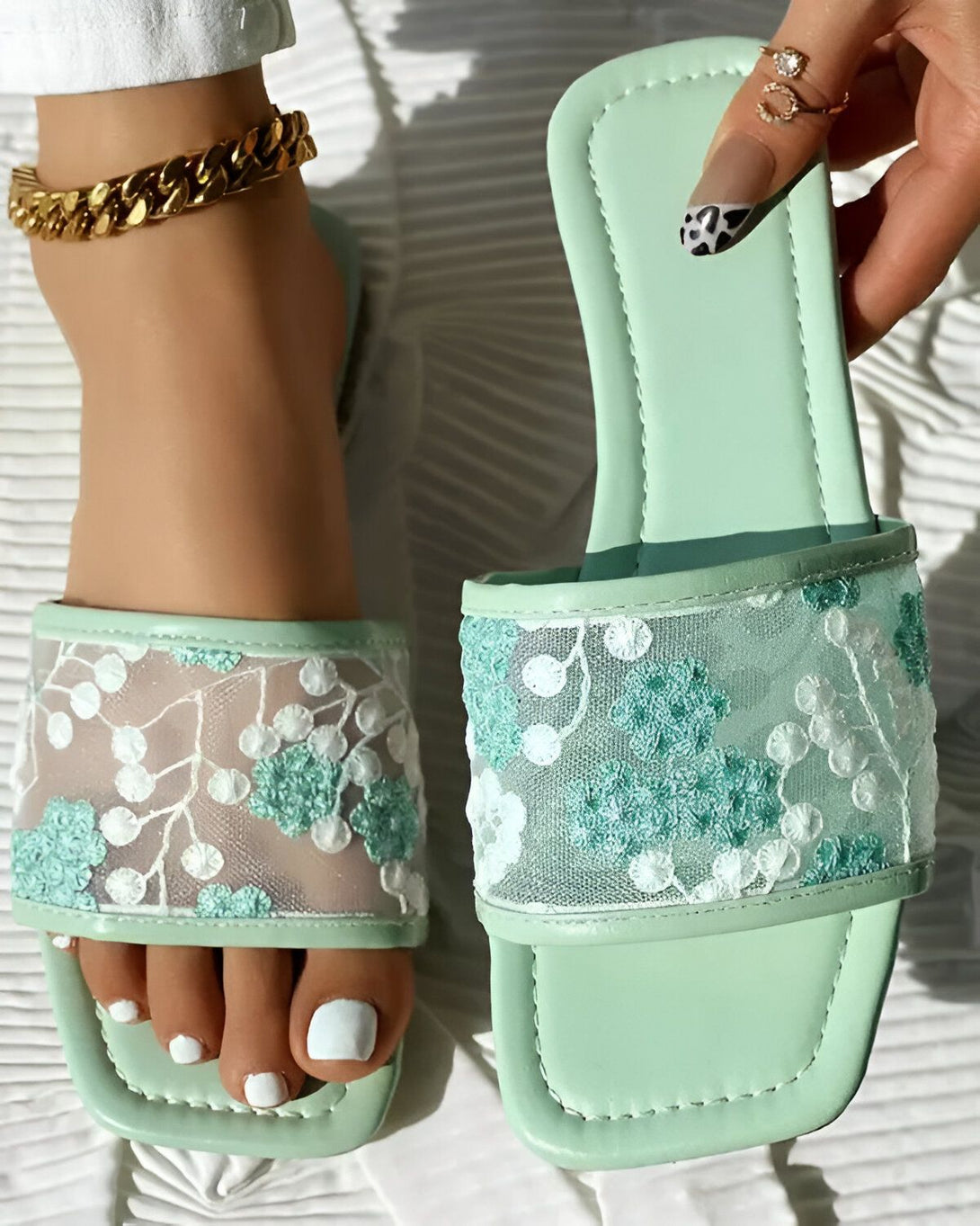 Une femme porte des sandales plates vertes à bout carré avec un motif floral brodé sur le dessus. Les sandales sont en similicuir et ont une semelle intérieure légèrement rembourrée. La femme a les ongles des pieds peints en blanc et porte un bracelet de cheville doré.