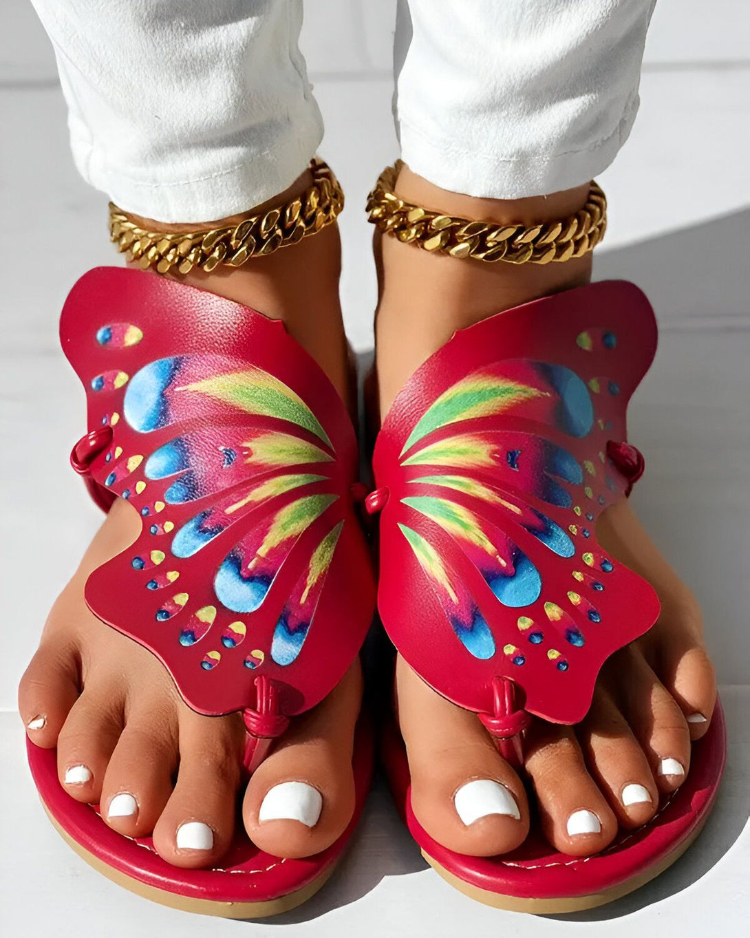 Une femme porte des sandales rouges en forme de papillon. Les sandales sont ornées d'un motif de papillons multicolores. La femme a les ongles des pieds peints en blanc et porte des bracelets de cheville en or.