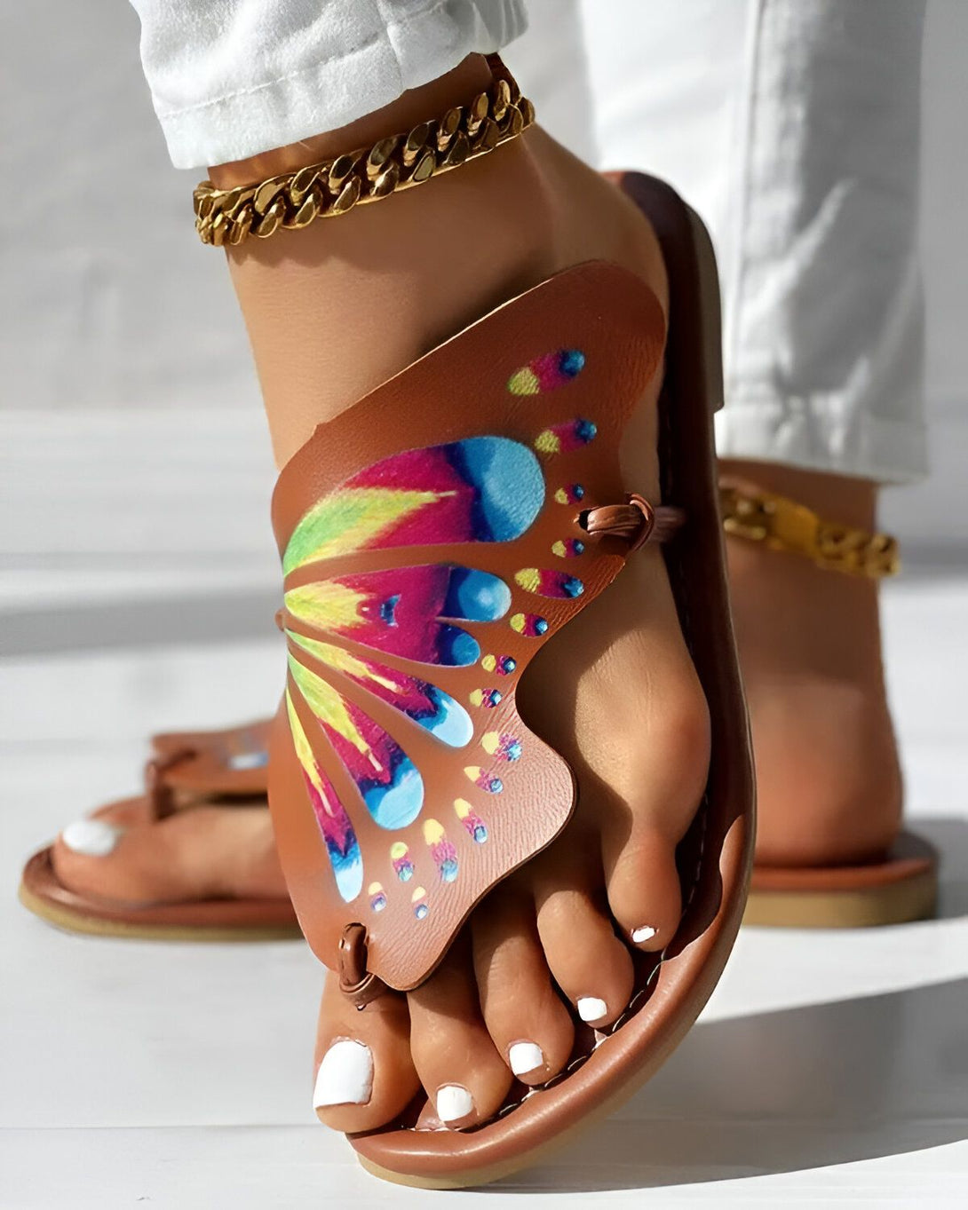 Une femme porte des sandales marron avec un motif papillon coloré. Les sandales sont en cuir et ont une semelle plate. La femme porte également un pantalon blanc et un bracelet de cheville doré.
