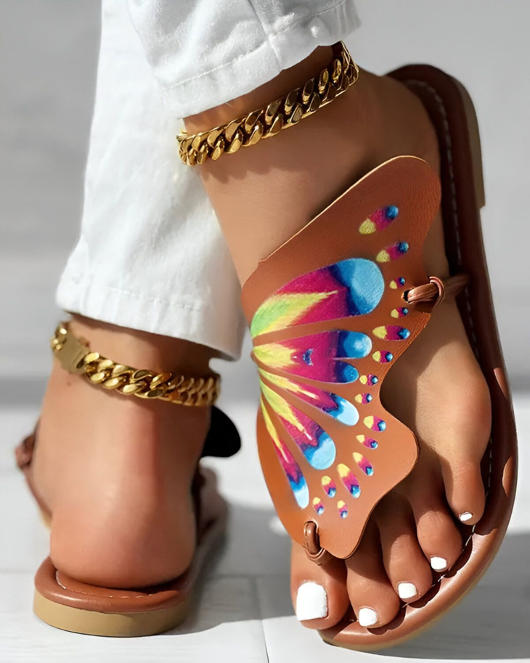 Une femme porte des sandales marron clair avec un motif papillon coloré. Elle porte également un pantalon blanc et des bijoux en or.