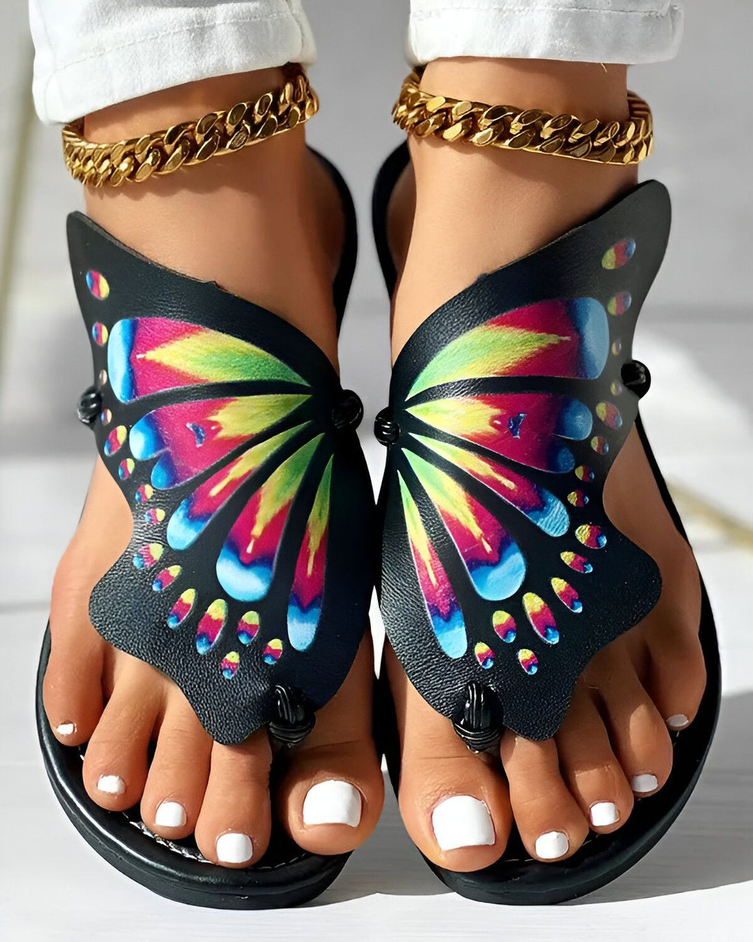 Une paire de sandales plates noires à brides croisées sur le dessus du pied. Les brides sont ornées d'un motif papillon coloré.