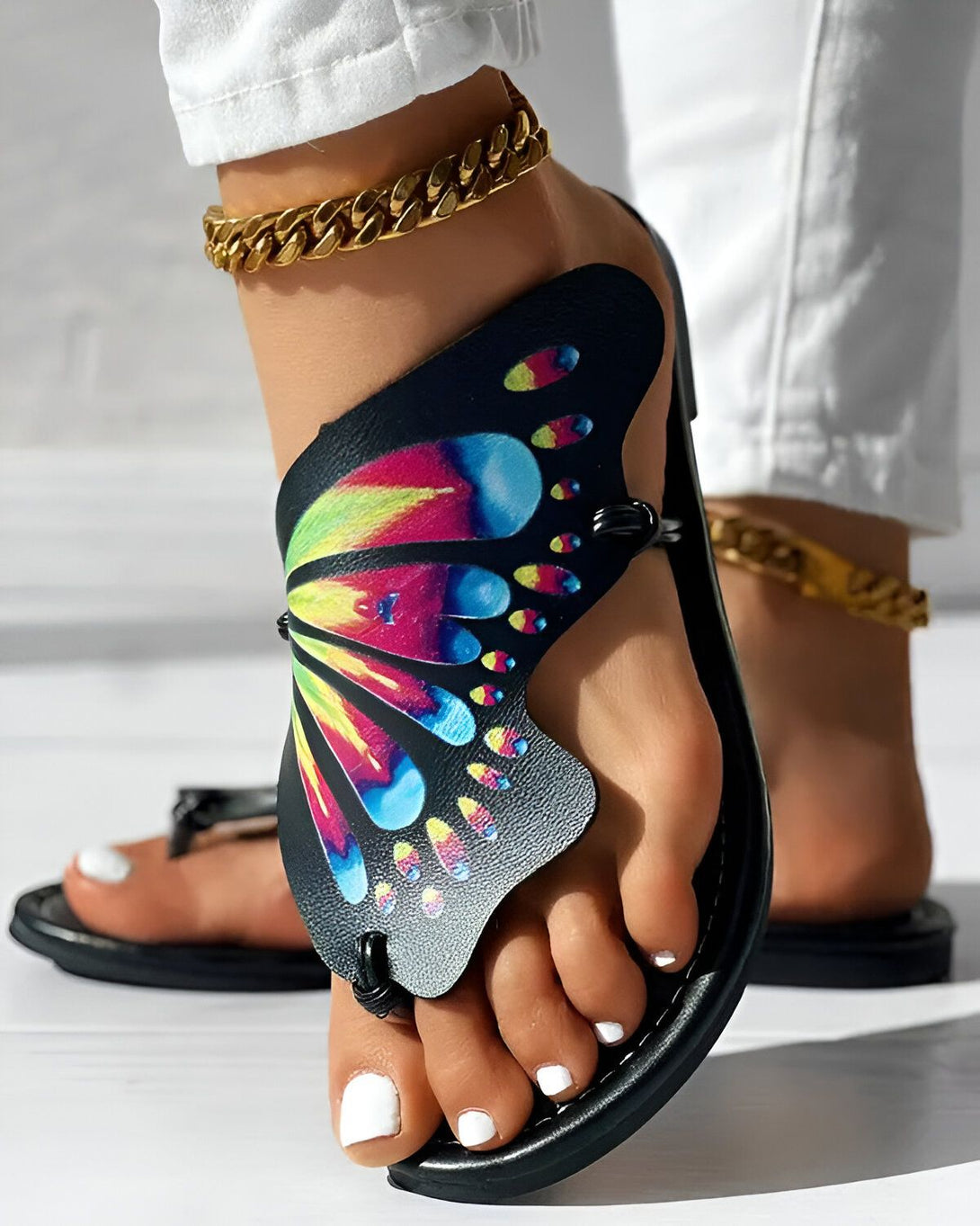 Une femme porte des sandales noires avec un motif de papillon coloré. Les sandales sont plates et ont une bride qui passe entre les orteils. La femme porte également un pantalon blanc et un chemisier blanc. Elle est debout sur un sol en bois.
