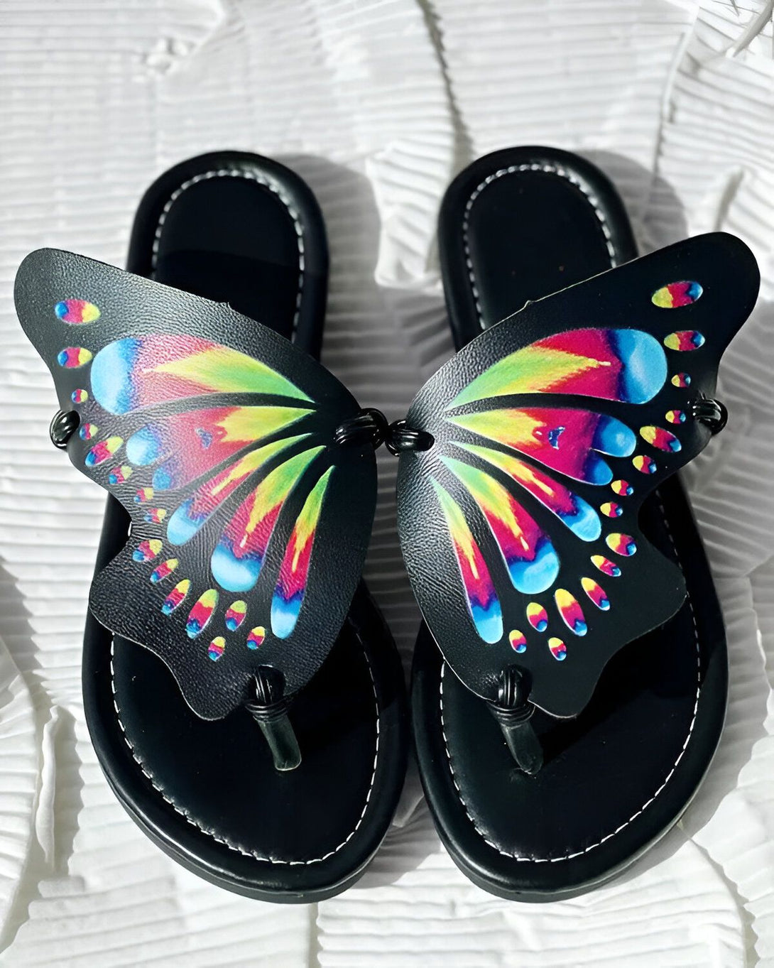 Une paire de tongs noires avec des ailes de papillon colorées. Les ailes sont faites de cuir et sont de couleur arc-en-ciel. Les tongs sont faites de caoutchouc et sont de couleur noire.