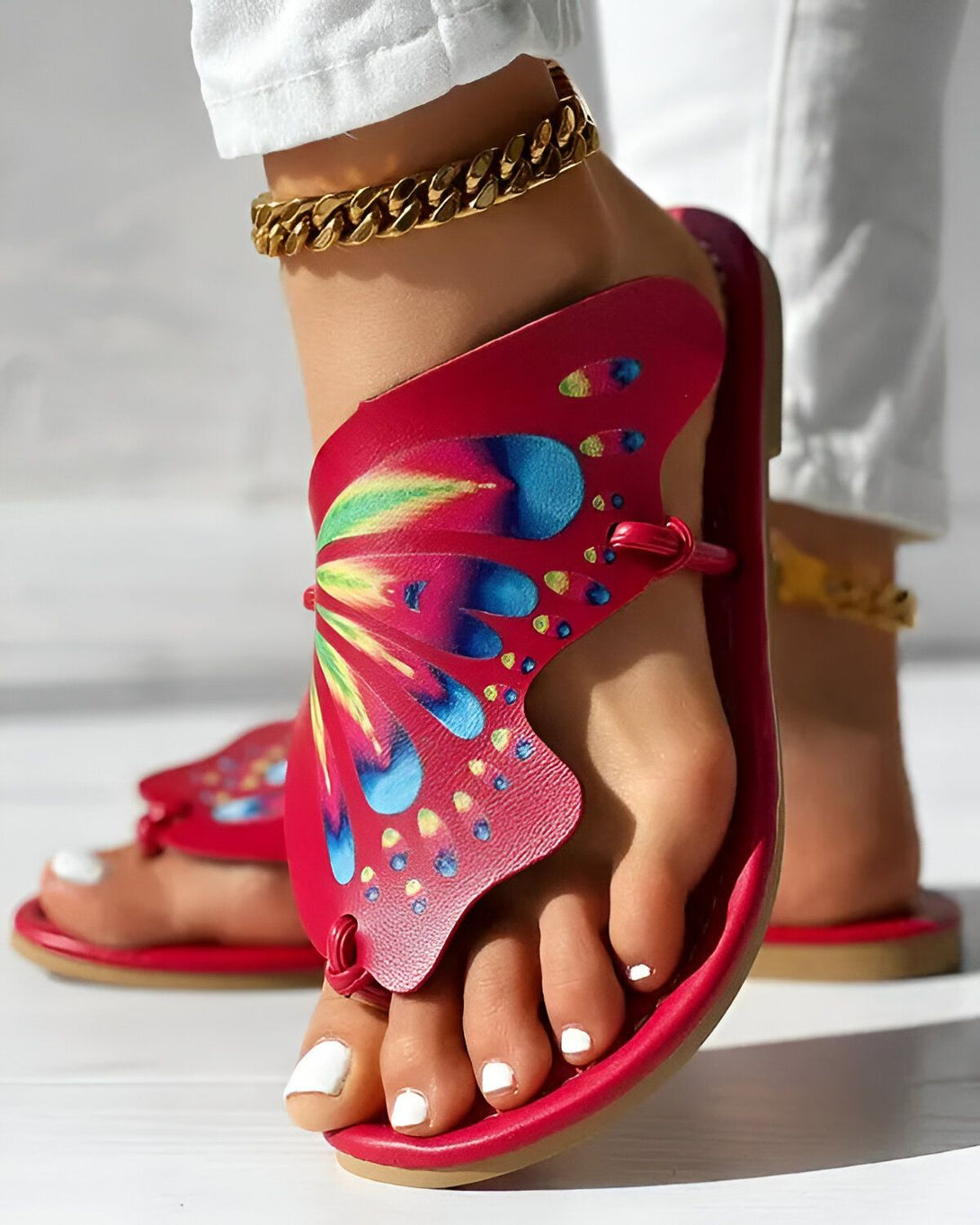 Une femme porte des sandales rouges avec un motif de papillon. Les sandales sont en cuir et ont une semelle plate. La femme porte également un pantalon blanc et un chemisier blanc. Elle a les cheveux longs et blonds et porte un collier doré.