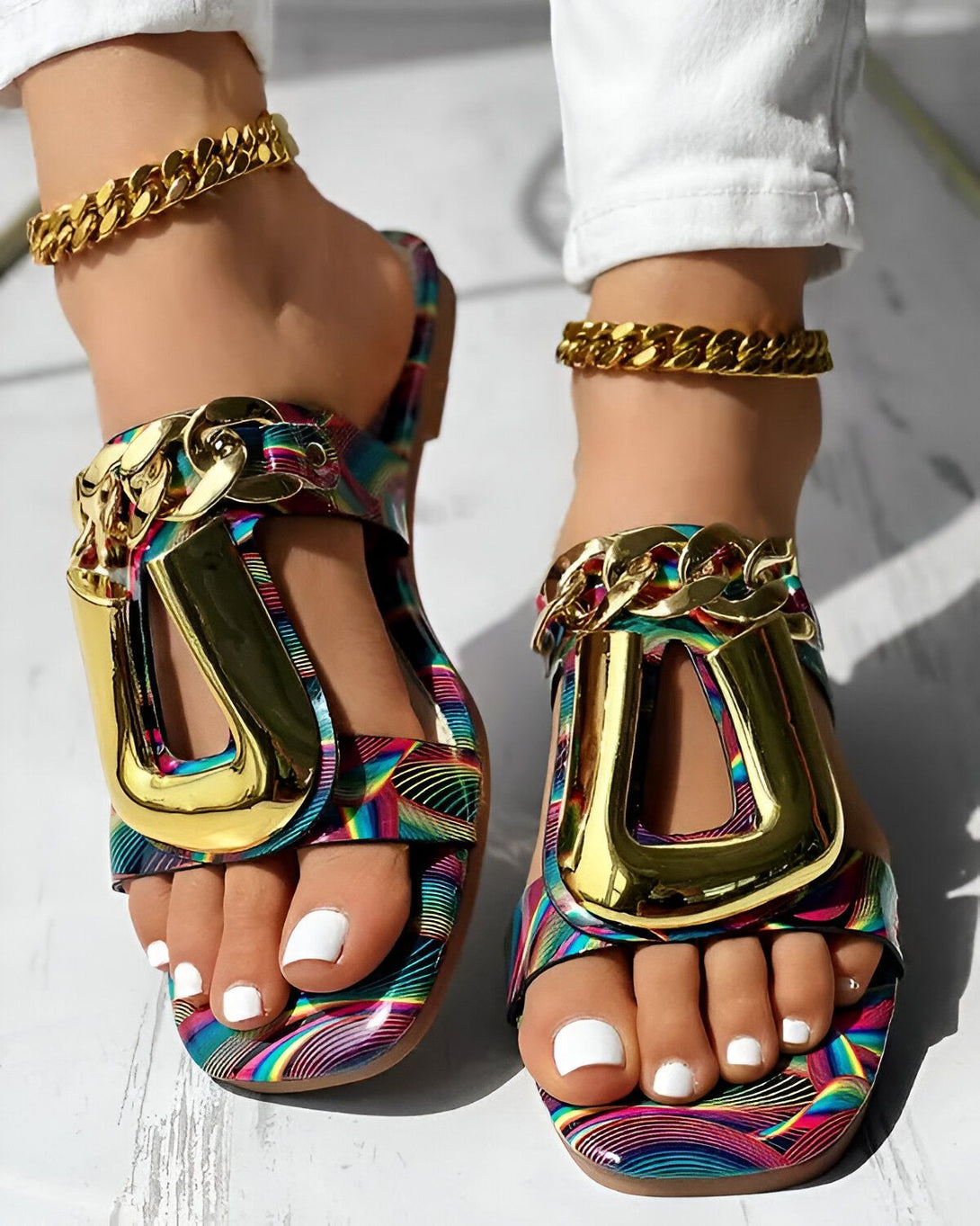 Une femme porte des sandales plates à brides larges et colorées. Les sandales sont ornées de chaînes dorées. La femme a les ongles des pieds peints en blanc.