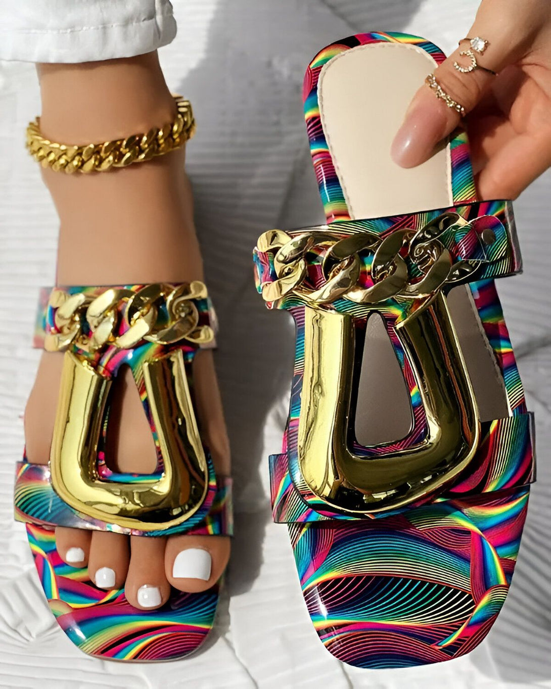 Une image de sandales plates à enfiler avec une bride sur le dessus du pied. Les sandales sont multicolores et la bride est dorée. Les sandales sont portées par une femme qui a les ongles des pieds peints en blanc.
