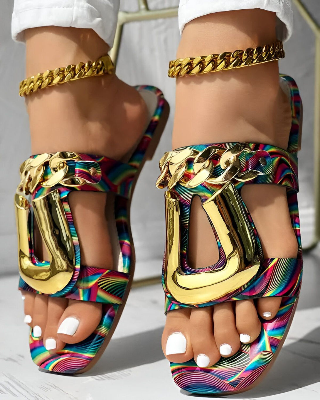 Une femme porte des sandales plates à brides dorées. Les sandales sont multicolores et ont un motif tie-dye. La femme a les ongles des pieds peints en blanc. Elle porte également des bracelets de cheville dorés.