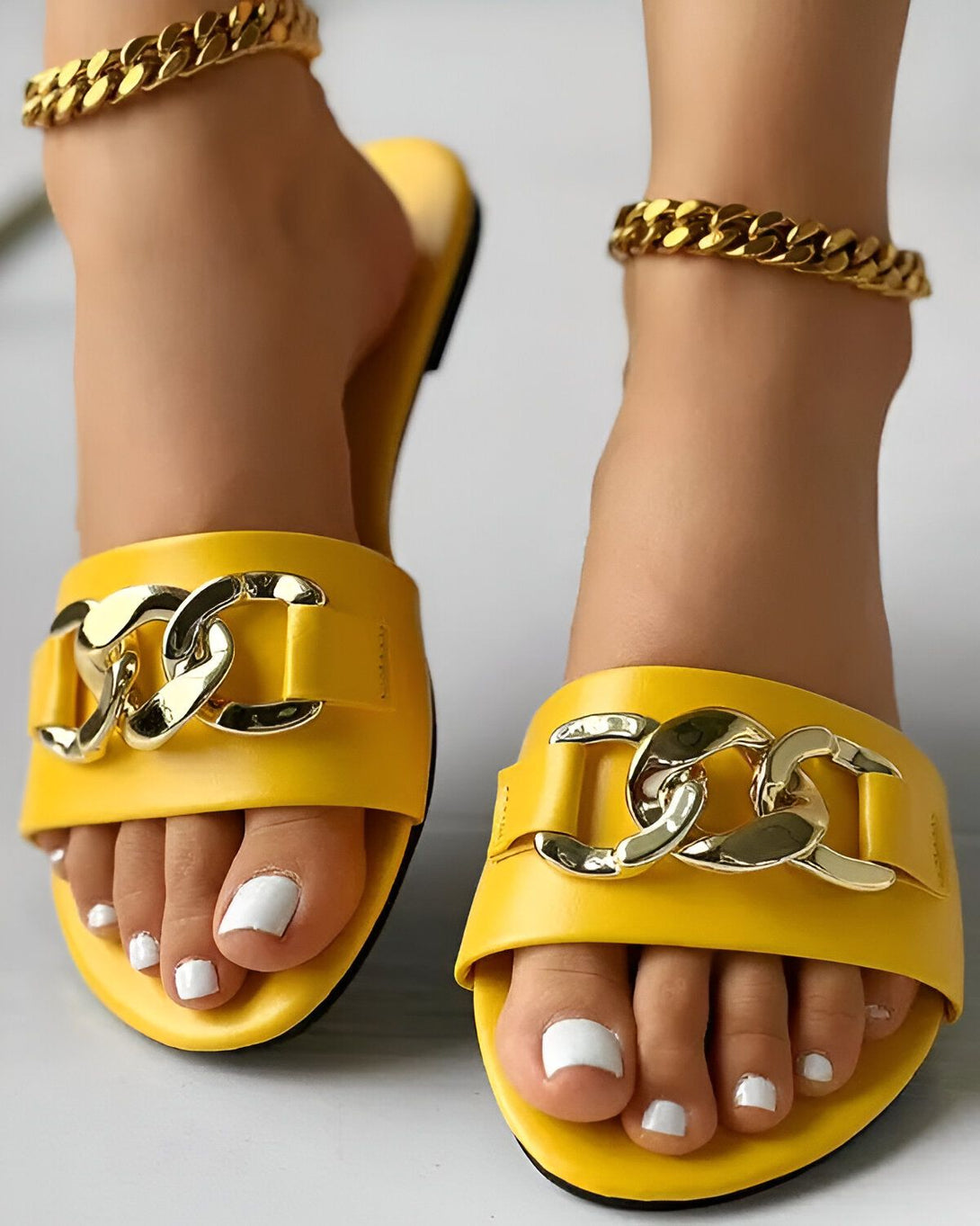 Une femme porte des sandales jaunes avec des chaînes dorées. Les ongles des pieds de la femme sont peints en blanc.