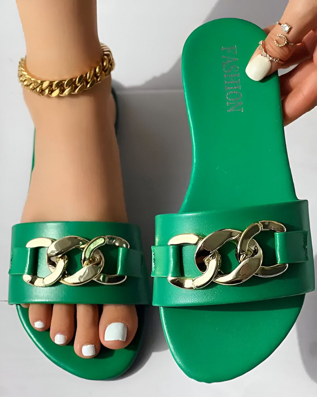 Une femme porte des sandales vertes avec des chaînes dorées. Les sandales sont plates et ont une bride qui passe entre les orteils. La femme a les ongles des pieds peints en blanc et porte un bracelet de cheville doré.