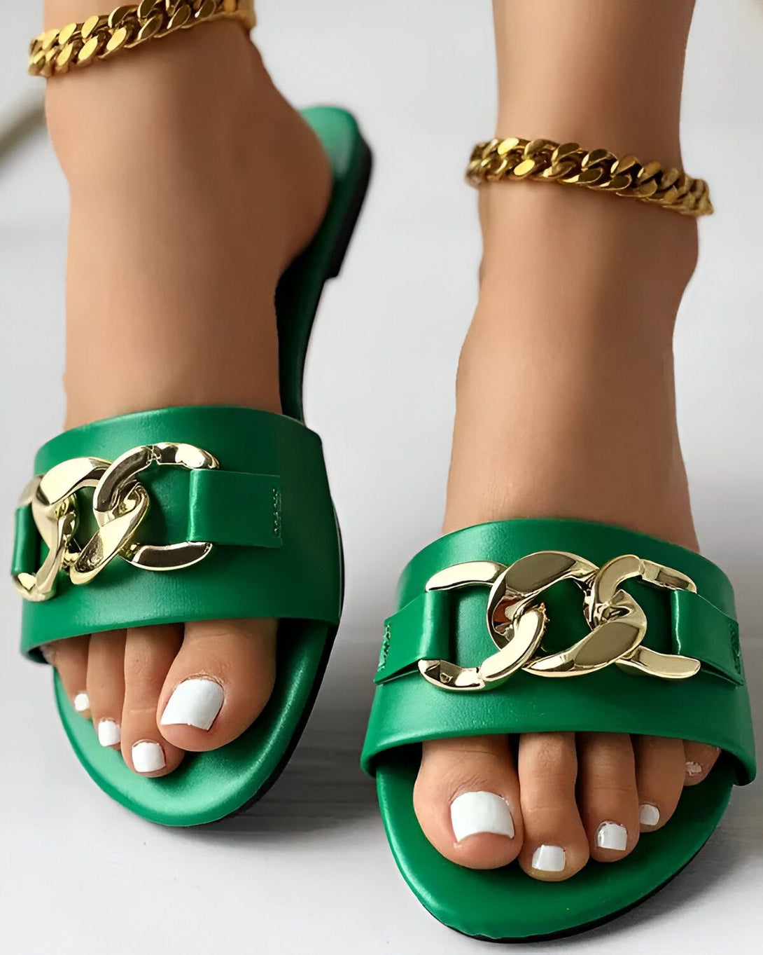 Une paire de sandales plates vertes avec une chaîne dorée sur le dessus. Les sandales sont portées par une femme avec une peau bronzée et des ongles blancs. La femme est debout sur un sol carrelé blanc.